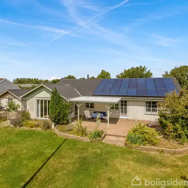 Huset har solpaneler på taget, to terrasser med havemøbler, omgivet af en velplejet have med buske og træer. Det ligger i et boligområde med lignende huse i baggrunden.