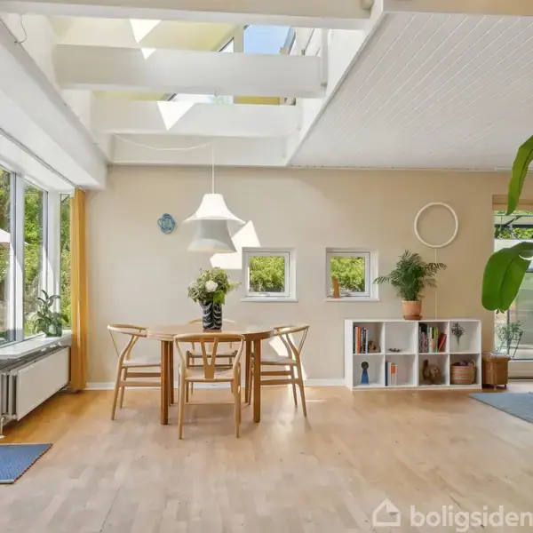 Et spisebord med fire stole står i et lyst rum med trægulv. Ovenlysvinduer giver naturligt lys, mens en hvid reol og grønne planter dekorerer rummet.