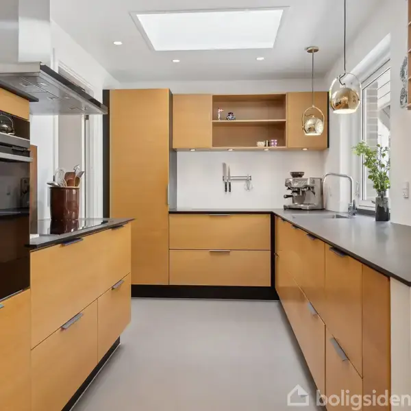 Køkken med træelementer, tilbyder indbyggede skabe og apparater, som ovn og kaffemaskine. Vinduer tillader naturligt lys, og åbne hylder har dekorationer. To hængende lamper skaber hyggelig belysning over arbejdsfladen.