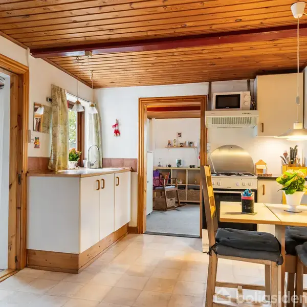 Køkken med lyst træloft og hvide skabe. Spisebord er dækket med blomsterpotte. Til højre, en åben dør viser en tilstødende stue. Rummeligt og lyst miljø.