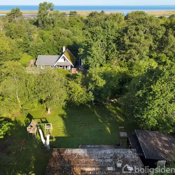 Et hus med gråt tag er omgivet af tæt skov. En legeplads med rutsjebane ses foran huset. I baggrunden er der udsigt til vandet.