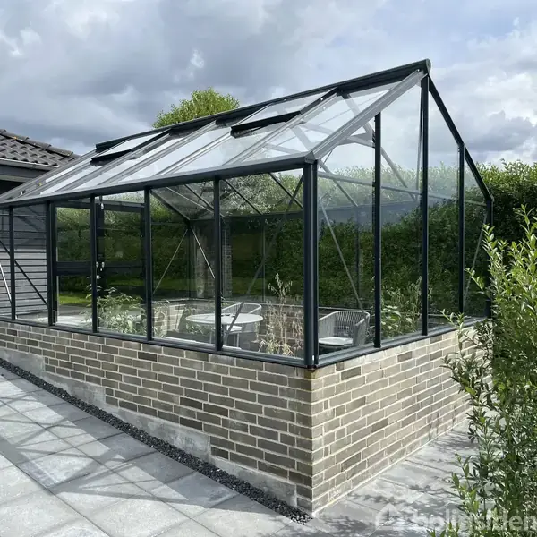 Drivhus med glaspaneler står på en lav murstensbase. Indeni findes stole og planter. Placeringen er ved siden af et hus med grå facade, omgivet af frodigt grønt og fliser.