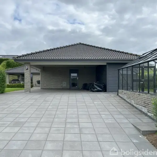 Carport står tom, flankeret af et drivhus til højre og grønne hække til venstre. Himmel er delvist overskyet, omgivelserne er velplejede og ordnede.