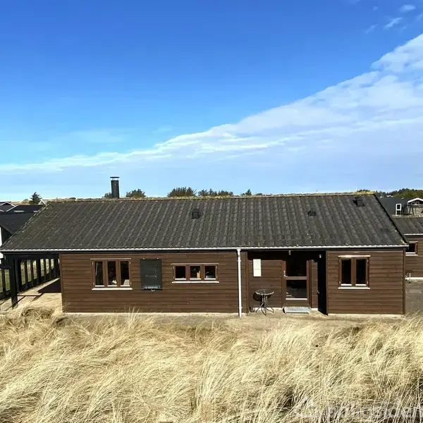 Et træhus står stille omgivet af høje klitter med strå. Huset har et sort tag og brune vægge, der omgiver en terrasse med et bord og to stole. Blå himmel i baggrunden.