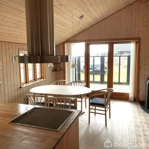 Køkken med kogeplade i forgrunden, spisebord med stole i midten og brændeovn ved siden af store vinduer, der fører ud til en grønt område. Trævægge og -loft.