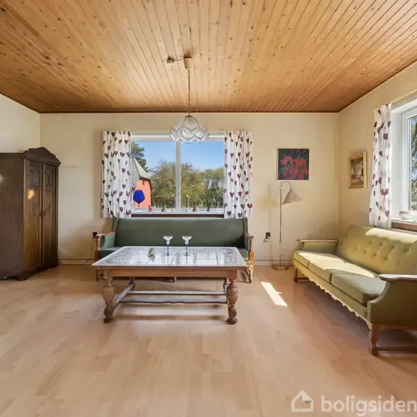 Stue med grøn sofa og træbord foran et vindue med blomstrede gardiner. En gammel skænk står ved siden af. Yderligere dekoration inkluderer en stålampe og maleri på væggen.