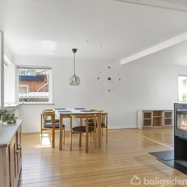 Spisebord med stole står i et lyst rum med hvide vægge og trægulv. Et ildsted er tændt i hjørnet, og der hænger en lampe over bordet.