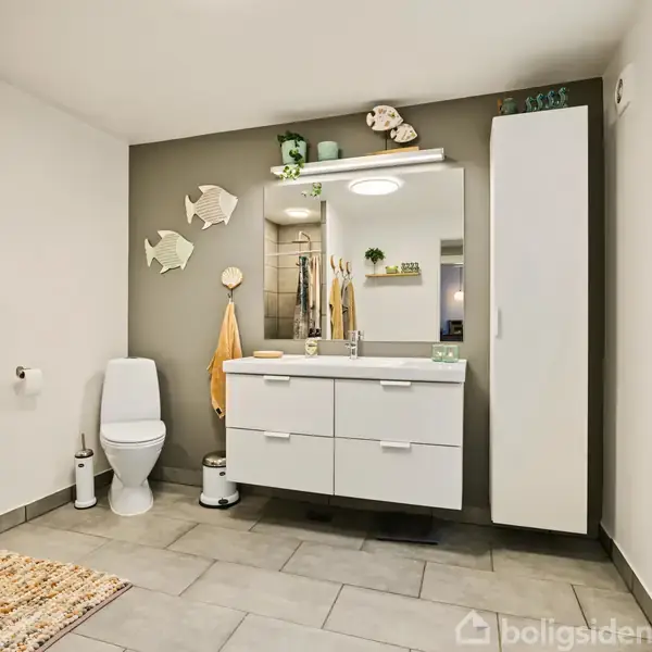 Toilet står ved siden af hvidt skab med vask og spejl. Håndklæder hænger på vægkrog. Dekorative fisk pryder bagvæggen, og gulvet er flisebelagt.