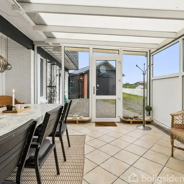 Et langbord med stole står i en lukket veranda med glasvægge. En enkelt stol i flet står ved siden af. Udendørs ses en have gennem glasdøren.