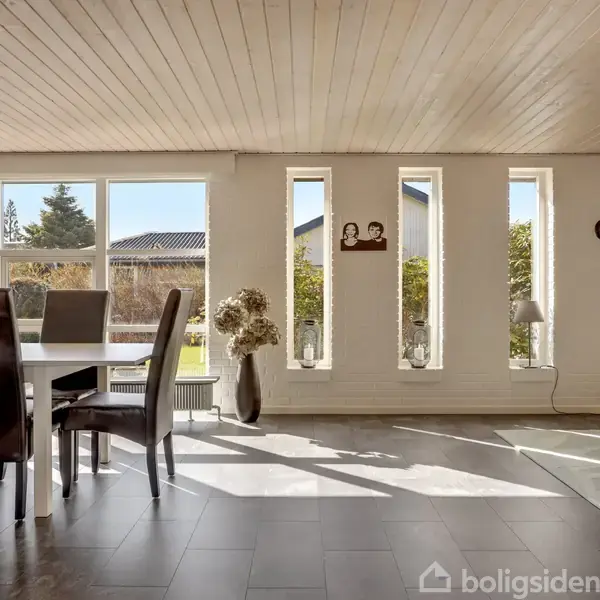 Spisestue med mørke stole omkring et bord. Sollys strømmer ind gennem store vinduer med udsigt til en have. Lyse trægulve og hvidmalet væg giver et indbydende rummiljø.