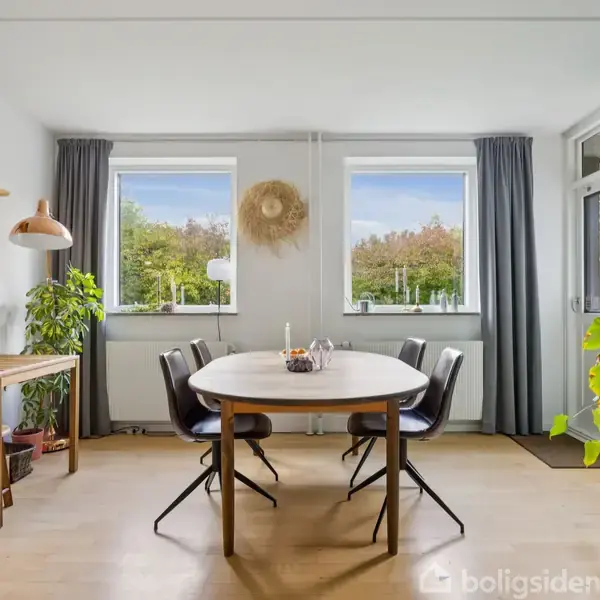 Et rundt spisebord med sorte stole står i en lys stue. To vinduer med grå gardiner bagved udsigt til træer. En blå væg til højre, planter og en reol til venstre.