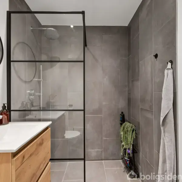 Et moderne badeværelse med en brusekabine med glasvægge til venstre, en træskab med håndvask, og en rund spejl. Grå fliser præger væggene. En håndklæde hænger på en krog.