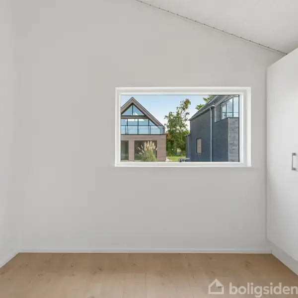 Et hvidt rum med trægulv indeholder et klædeskab til højre. Gennem vinduet ses moderne huse og grønne træer udenfor.