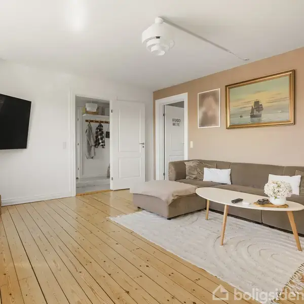 Brun sofa står på lyse trægulve, placeret foran et rundt sofabord og et vægmonteret TV. Baggrunden inkluderer et åben døråbning og vægkunst.
