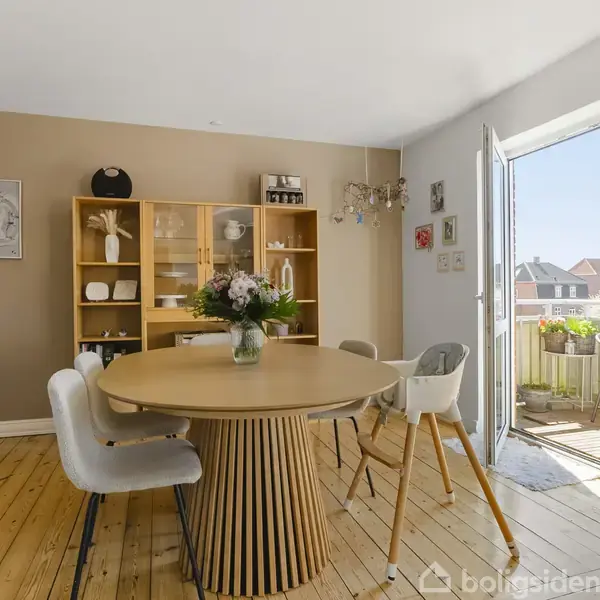 Et rundt spisebord står midt i en lys spisestue med åbne døre til en balkon. I baggrunden er der en reol med dekorationer og vægkunst. Ingen tekst.