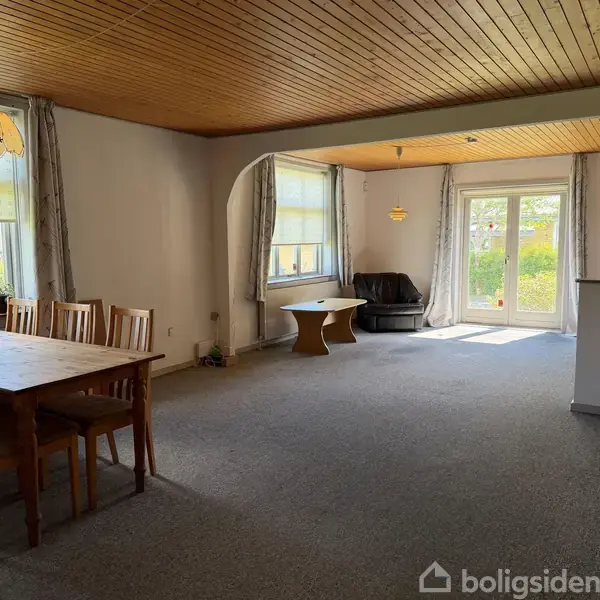 Spisestuebord med stole står i en stue med tæppebelagt gulv. I baggrunden ses et sofamøbel nær et vindue. Rummet har lyst træloft og store vinduer med gardiner.