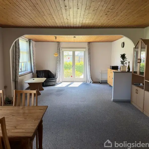 Stue med trægulv og træloft, spisebord i forgrunden, sofa ved vindue mod haven i baggrunden, og skabsarrangement til højre. Lys strømmer ind fra glasdør.