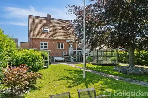 Murstenshus med rødt tag står i solen. En grussti fører gennem en frodig have med græs, træer og buske. Der er havesæt og en flagstang i baggrunden.