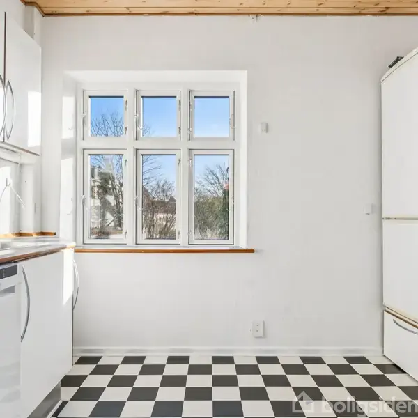 Et lyst køkken med sort-hvidt ternet gulv. Hvide skabe og apparater står langs væggen. Et vindue i midten giver udsigt til træer udenfor, med blå himmel.