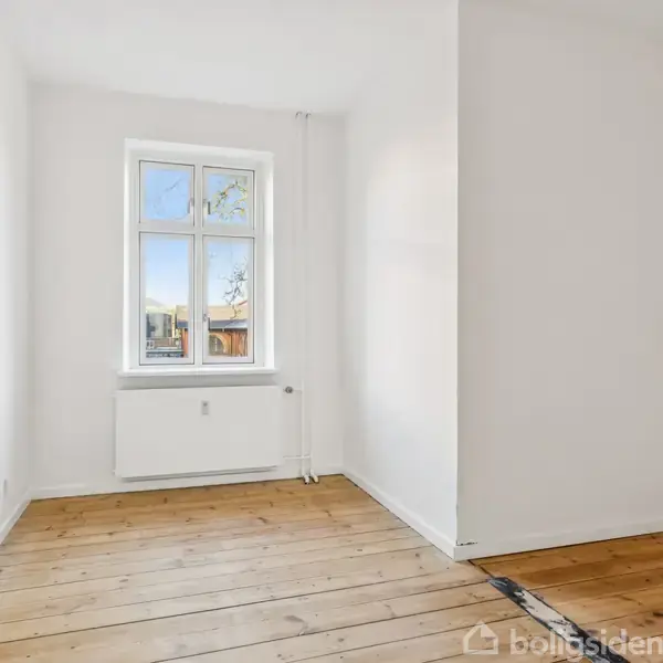 Værelse med lyse trægulve har sparsom indretning. Vinduet viser udsigt til et solrigt udendørsområde. Væggene er hvide, og der er en radiator under vinduet.