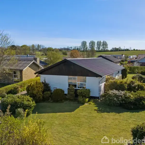 En enkeltstående hvid bygning med mørkt tag er omgivet af en velplejet have med buske. Andre huse og træer er synlige i baggrunden under en klar blå himmel.