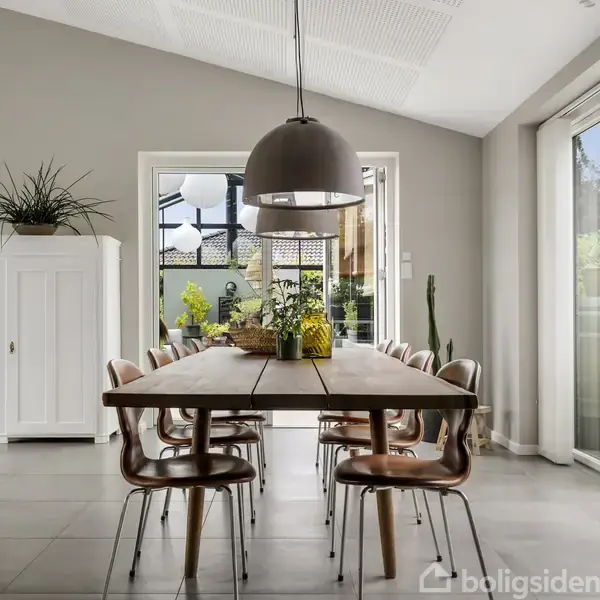 Spisebord med seks stole står i et lyst rum med grå vægge. En stor lampe hænger over bordet. Rummet har store vinduer med udsigt til en terrasse med planter.
