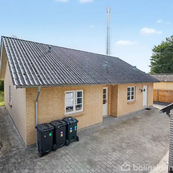 Hus med gult murværk og skråt tag; står i en flisebelagt gårdsplads med affaldscontainere foran, omgivet af træer og hegn.