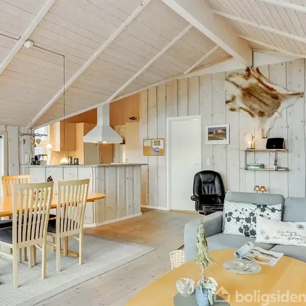 Stue med spisebord og seks stole, en sofa, brændeovn og åben adgang til køkken. Trævægge og synlige loftsbjælker skaber en hyggelig atmosfære. En bjørneskind dekoration hænger på væggen.