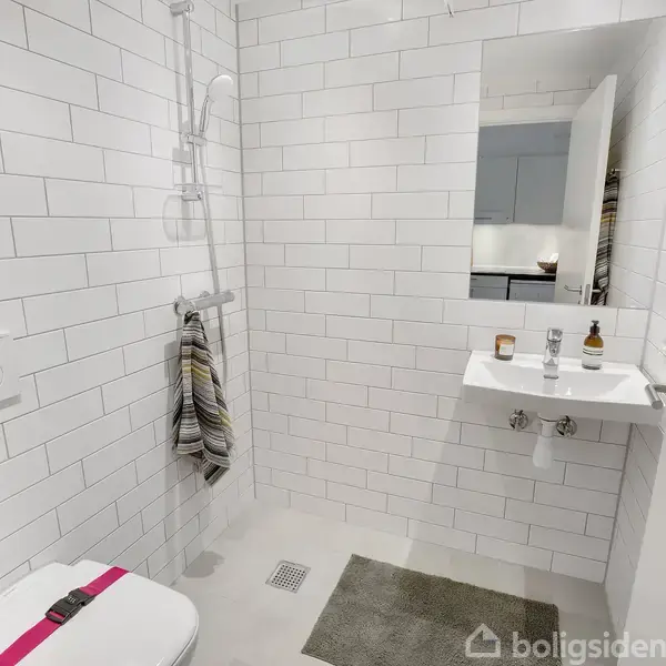 Et moderne badeværelse har en hvid flisebelægning på vægge og gulv. Et toilet sidder til venstre, en bruser med hængende håndklæde i midten og en vask med spejl til højre.