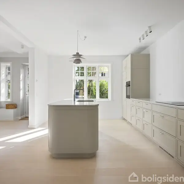 Køkkenø står i et lyst, minimalistisk køkken med lyse trægulve. Hvide skabe fylder højre side. Store vinduer bader rummet i naturligt lys. Lampen hænger over øen.