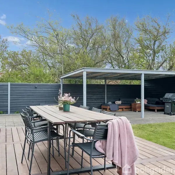 Et træbord med stole i en havepavillon. En lyserød plaid hænger over en stol. I baggrunden ses en grill under en overdækket terrasse ved et højt, gråt træhegn.