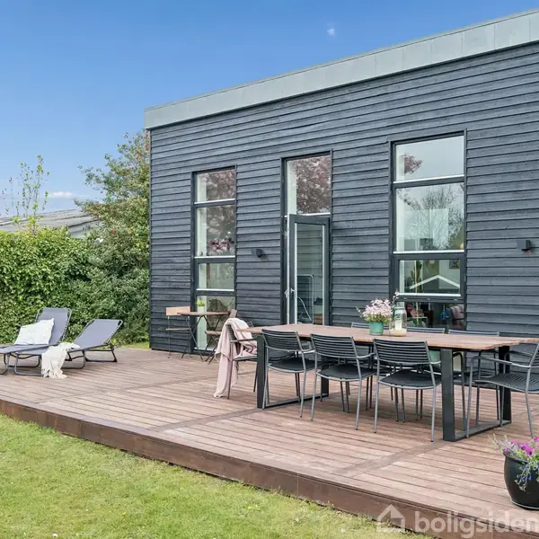 En moderne terrasse har et langbord med stole, omgivet af en græsplæne og buske. Ved siden af står to liggestole. Bygningen har sort træbeklædning og store vinduer.