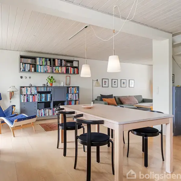 En lys spisestue med et træbord omkranset af sorte stole. Til venstre står en blå lænestol ved siden af en reol fyldt med bøger, og en sofa i baggrunden. En trappe fører opad.