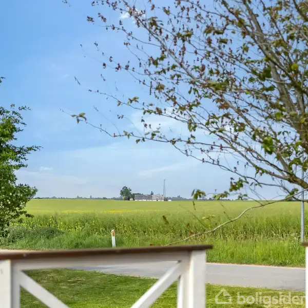 Terrasse med hvidt gelænder rammer en grøn mark i en landlig scene. Træer omkranser billedet, og himlen er blå. En stenet vej ses i baggrunden.