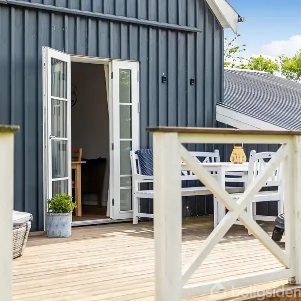 Terrasse med træhegn og åbne franske døre fører ind til et hus med en mørkeblå facade. Haven og grønne træer er synlige i baggrunden.
