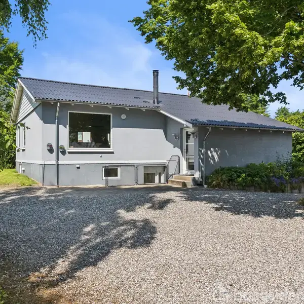 Et gråt hus står stille omkranset af buske og træer, med en stor grusvej foran. Blå himmel og sollys skaber en fredelig atmosfære.