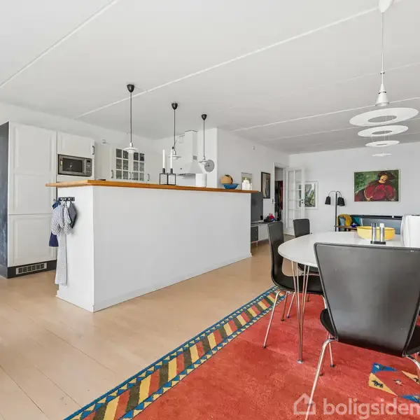 Spisebord med sorte stole står i køkken-alrum, omgivet af lyse trægulve og moderne indretning, med malerier på væggen og hvide skabe langs køkkenområdet.