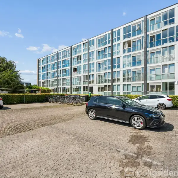 Flere biler holder parkeret på en asfalteret plads foran en moderne, fler-etagers bygning med mange vinduer. I baggrunden er der grøn beplantning og en blå himmel.