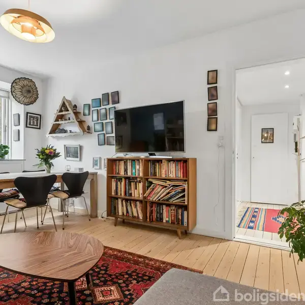 Stue med trægulv, rød orientalsk tæppe, sofa, rektangulært spisebord og sorte stole. Vægmonteret TV over en bogreol. Vindue med gardiner til venstre. Planter og diverse dekorationer.
