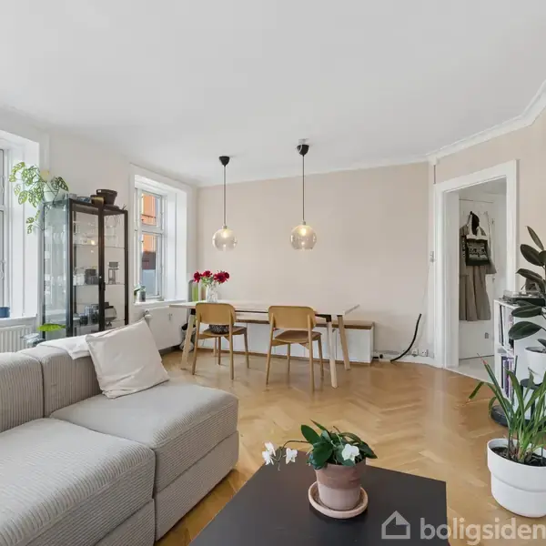 En lys stue med en grå sofa og sofabord i forgrunden. Spisebord med stole og hængende lamper i baggrunden. Vinduer slipper dagslys ind, og planter dekorerer rummet.