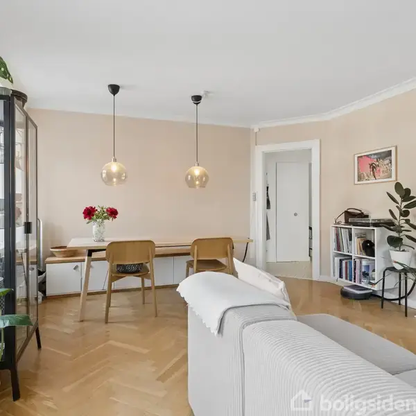 En stue med et gråt sofaarrangement, spisebord med to stole, omkranset af planter og reoler. To hængelamper over bordet. Maleri og åbne døre i baggrunden, parketgulv.
