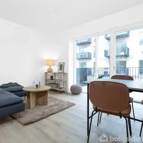 Stue med en blå sofa og sofabord på en lys tæppe. Et spisebord med brune stole står ved siden af store vinduer med udsigt til altan og bygninger udenfor.