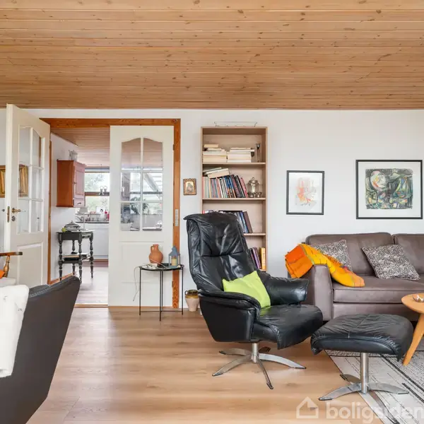 En stue med en sort lænestol og fodskammel i centrum. En grå sofa med farvede puder står til højre. Omgivelserne har trægulve, loft i træ og lys indretning.
