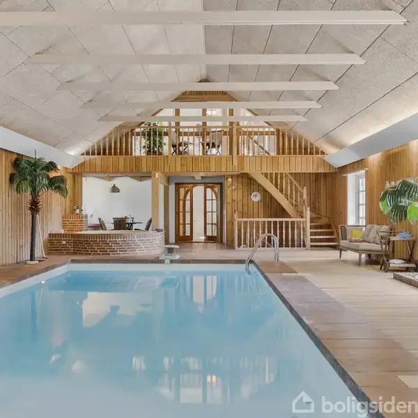 Indendørs swimmingpool i et rum med trævægge og loftsbjælker. Poolen fylder det meste af gulvet. Der er planter, siddepladser og en trappe til en lille hems i baggrunden.