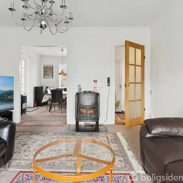 En stue med sorte lædersofaer omkring et rundt glasbord på et flerfarvet tæppe. En brændeovn står ved en hvid væg nær en åben dør til en spisestue. En lysekrone hænger i loftet.