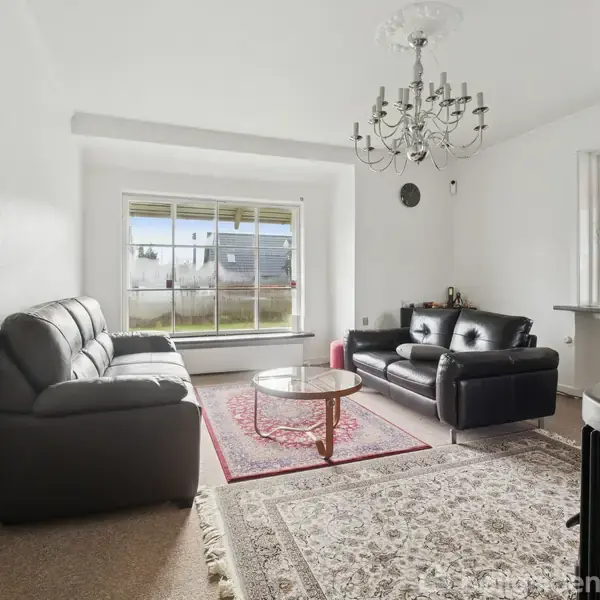 To sorte lædersofaer står i en stue med lysindfald fra store vinduer og farverig glasvæg. En lysekrone hænger over et rundt sofabord på et persisk tæppe.