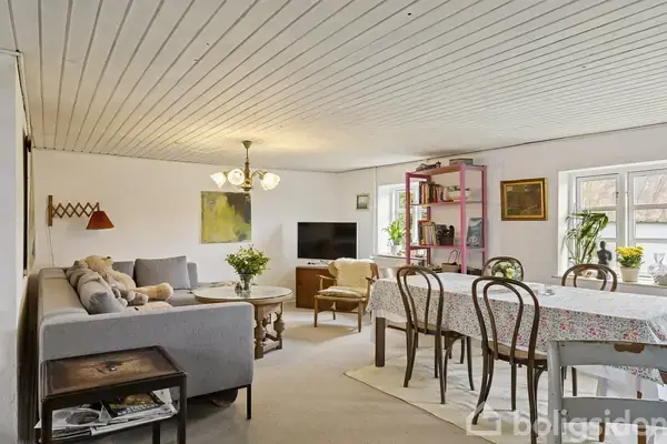 Gråt sofaarrangement med puder og bamser, placeret i en lys stue. Rundt bord foran sofaen, spisebord med stole i nærheden, omgivet af hvide vægge og vinduer.