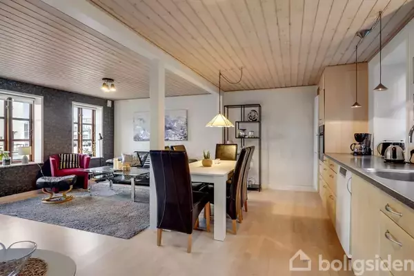 Stue og køkken i åbent rum; spisebord med seks stole står centralt, omgivet af moderne møbler og køkkenindretning. Store vinduer og lys trægulv skaber en lys og indbydende atmosfære.