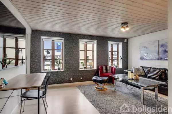 En stue indeholder en stor sofa med puder og et glasbord. En rød lænestol står nær tre vinduer, der lukker naturligt lys ind. Baggrunden viser en grå væg.