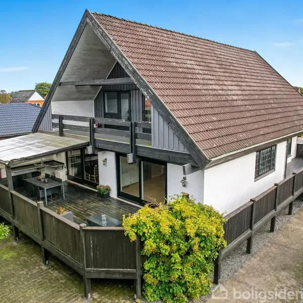 Huset med skråt tag og balkon står i et boligområde. Der er en overdækket terrasse i en indhegnet have med buskagtige planter langs hegnet.